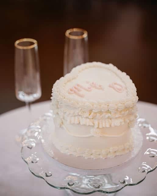 A small, heart-shaped white cake with decorative icing sits on a glass plate next to two empty champagne flutes on a round white tablecloth.