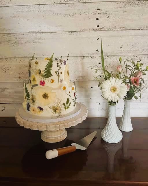 A two-tier cake decorated with pressed flowers sits on a wooden cake stand next to two white vases holding fresh flowers, all placed on a dark wooden table against a rustic white plank wall. A cake server lies in front.