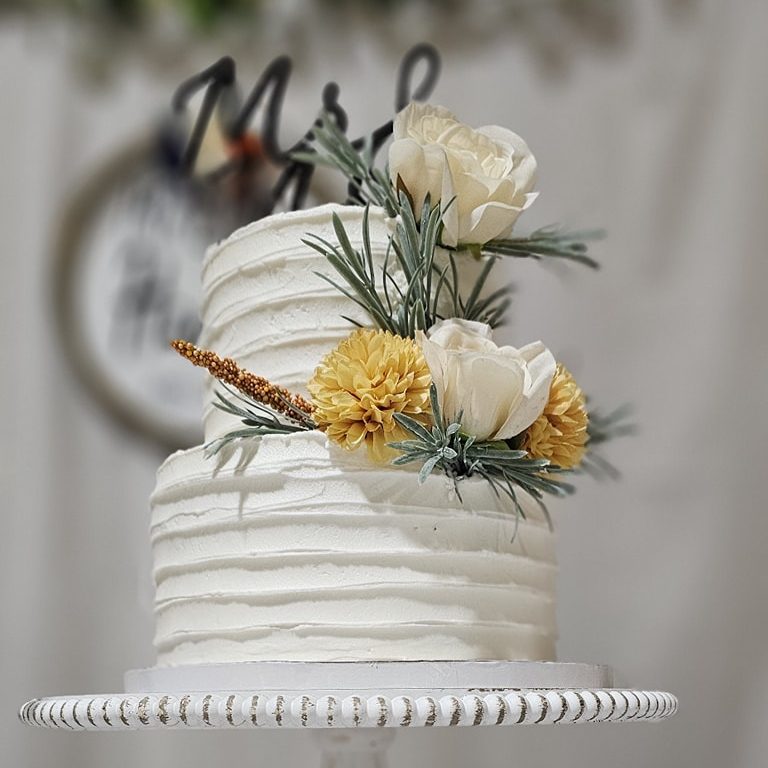 A two-tiered white cake with textured frosting, decorated with white and yellow flowers and greenery, sits on a white cake stand. The background is blurred with some green and white decor visible.